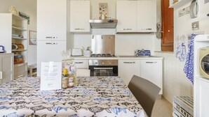 Eine kleine Küche mit weißen Schränken, einem Gasherd, einem Backofen und einem Kühlschrank. Ein Esstisch mit gemustertem Tischläufer und ein Stuhl sind vorhanden. Dekorative Elemente und ein sternförmiges Wandbild sind sichtbar.