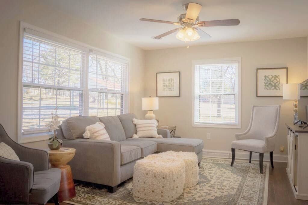 Un salon avec un canapé gris, un fauteuil blanc, un ventilateur de plafond et de grandes fenêtres équipées de stores.