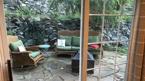 Een terras met rieten meubilair, een stenen muur en uitzicht door een glazen deur.