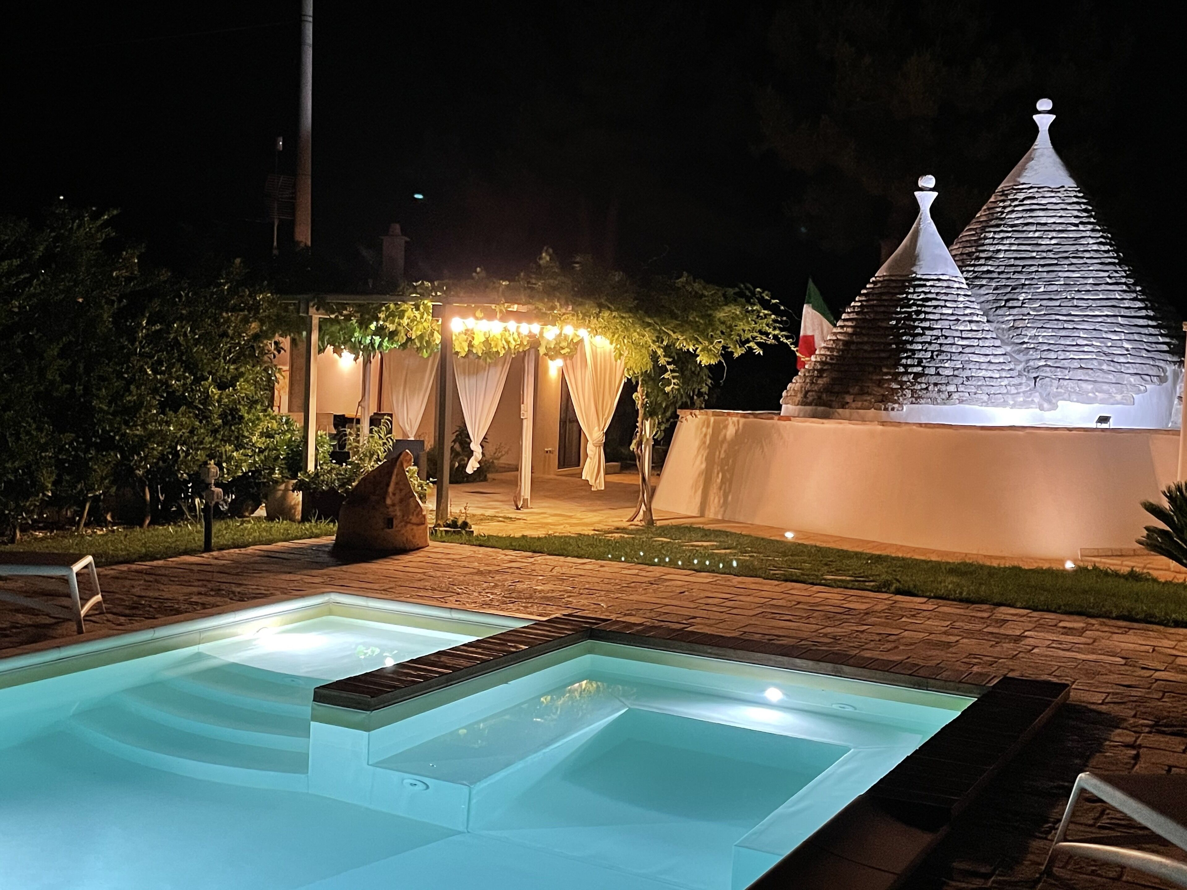 A nighttime pool area with a clear water surface, a stone structure with a pointed roof, and a covered seating area with string lights.