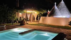 Ein Poolbereich bei Nacht mit ruhiger Wasseroberfläche, einer Steinstruktur mit spitzer Dachform und einer überdachten Sitzgelegenheit mit Lichterkette.