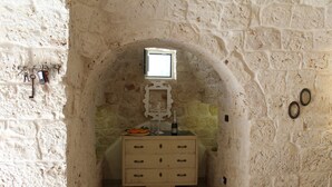 Ein schmaler Raum mit Steinwänden, einem Holzschrank, einem kleinen Fenster und einem an der Wand befestigten Regal mit Schlüsseln und einer Flasche.