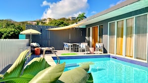 Ein Swimmingpool mit Liegestühlen und einem Tisch, umgeben von einer Hecke und Grünpflanzen.