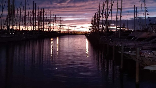 Ein Yachthafen mit mehreren vertäuten Booten, ruhigem Wasserspiegel, der die Boote reflektiert, und einem Sonnenuntergang im Hintergrund.