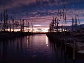 A marina with multiple boats docked, calm water reflecting the boats, and a sunset in the background.