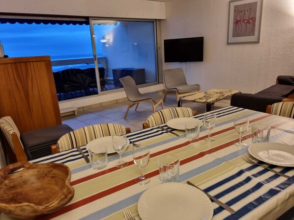 A dining area with a table set for four, a television mounted on the wall, and a view of the sea through a glass door.