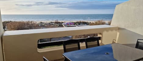 A balcony with a table and chairs, overlooking a beach with a striped umbrella.