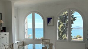 Un espace repas avec une table en verre et des chaises blanches, de grandes fenêtres en arc de cercle offrant une vue sur la mer, et un ventilateur de plafond.