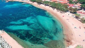 Vue aérienne d’une zone côtière avec des eaux turquoise limpides, une plage de sable et des immeubles résidentiels aux toits rouges.