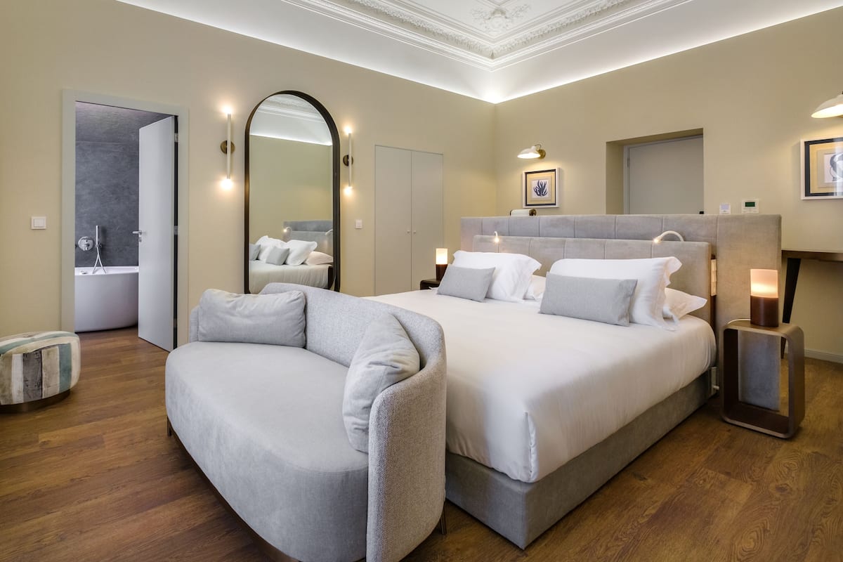 Ein modernes Hotelzimmer mit einem großen Bett, einer Couch, einem Spiegel und einem durchgegangene Tür sichtbaren Badezimmer.
