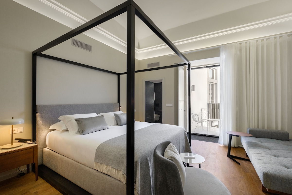 A four-poster bed with a grey headboard, a bedside table with a lamp, and a grey sofa in a modern hotel room.