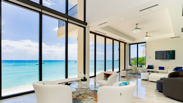 A spacious living room with a large window overlooking a beach, furnished with white sofas and a glass coffee table. - Oasis 7-stunning private beachfront villa with rooftop pool-by Cayman Villas! (Seven Mile Beach)