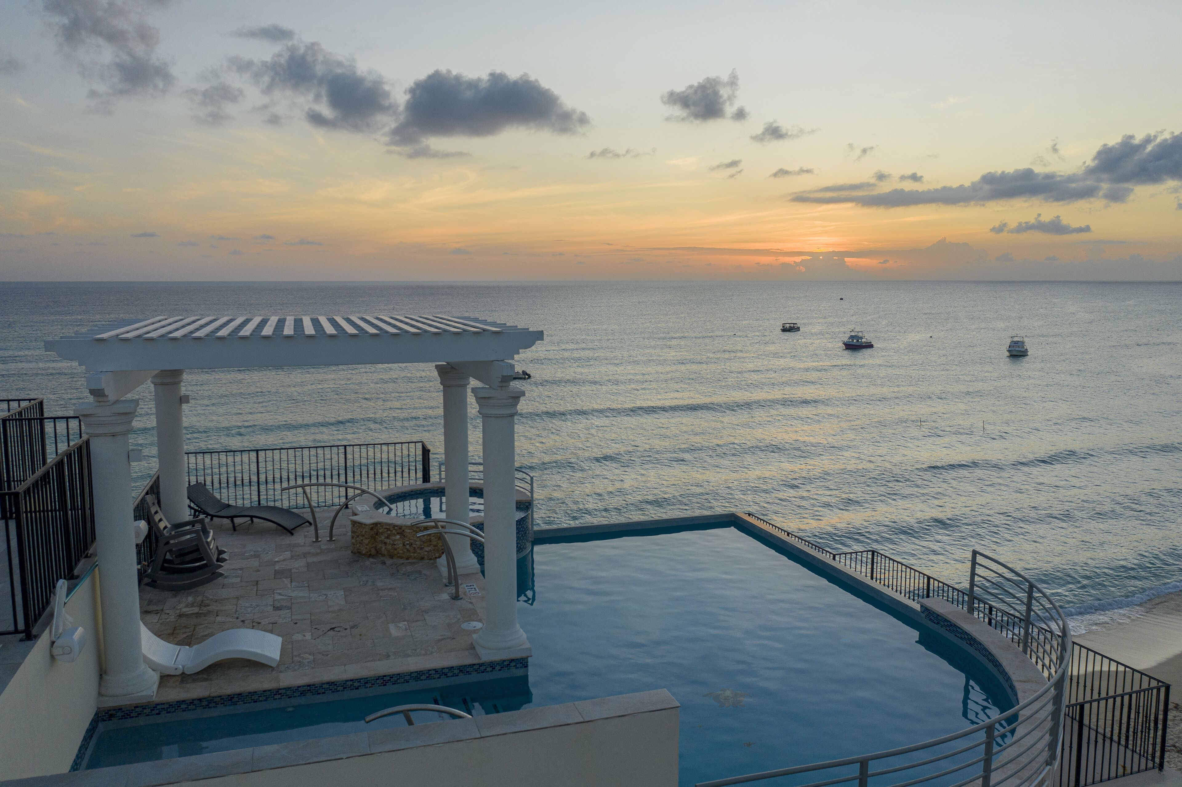 A poolside area with a pergola, lounge chairs, and a view of the sea at sunset.
