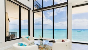 A spacious room with a large balcony overlooking the ocean, featuring white furniture and a glass coffee table. - Oasis 7-stunning private beachfront villa with rooftop pool-by Cayman Villas! (Seven Mile Beach)