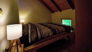 Et soveværelse med skrå væg, seng med mønstret dyne, sengetåget lampe og et vindue med udsigt til grønne omgivelser.