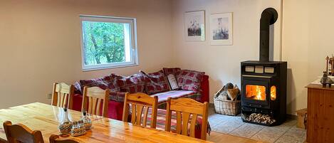 Een gezellige woonkamer met een houten tafel en stoelen, een bank, een open haard en een raam met uitzicht op bomen.