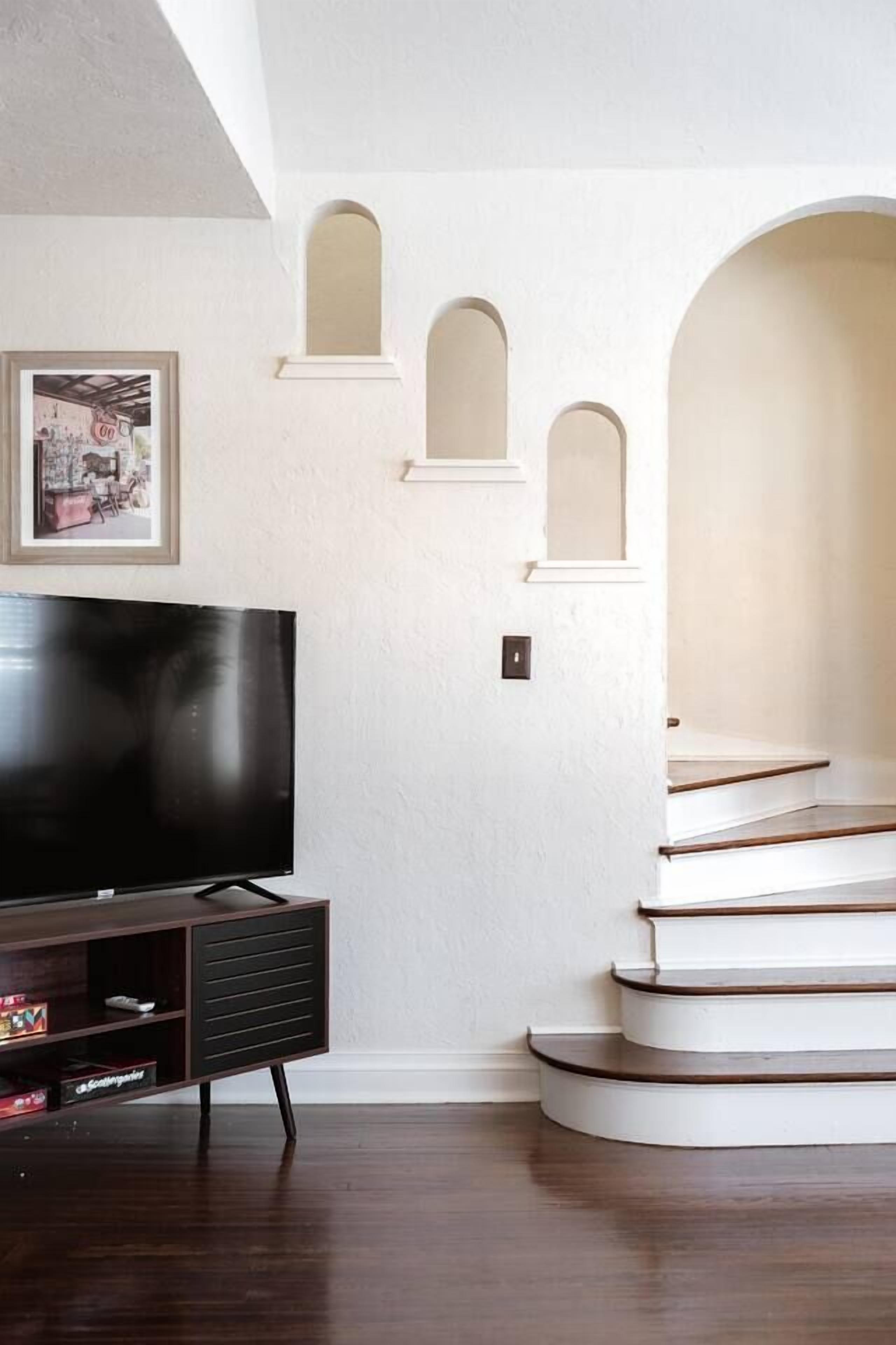 Ein modernes Wohnzimmer mit einem Flachbildfernseher auf einem dunklen Holzschrank, weißen Wänden und einer Treppe mit weißen Stufen.