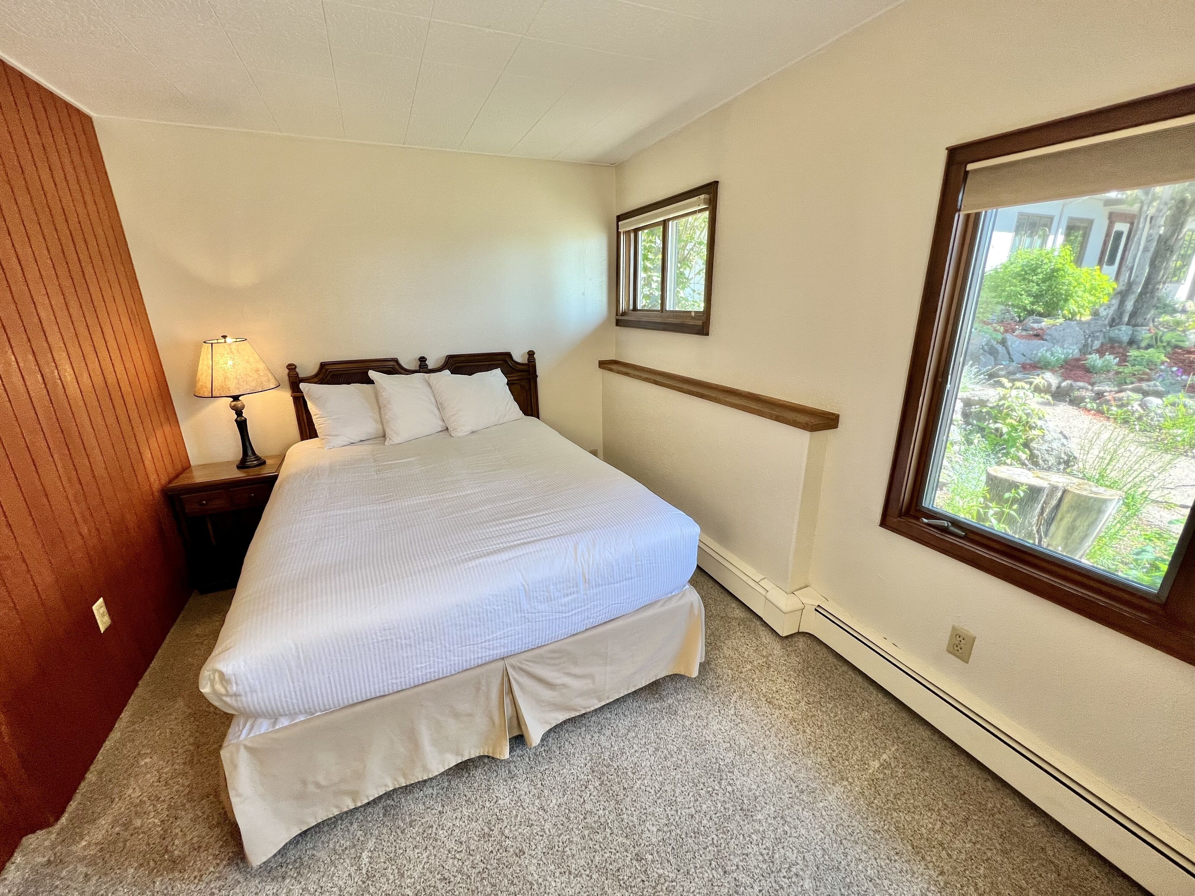 A neatly made bed with white linens, a bedside lamp, and a window with a view of greenery.