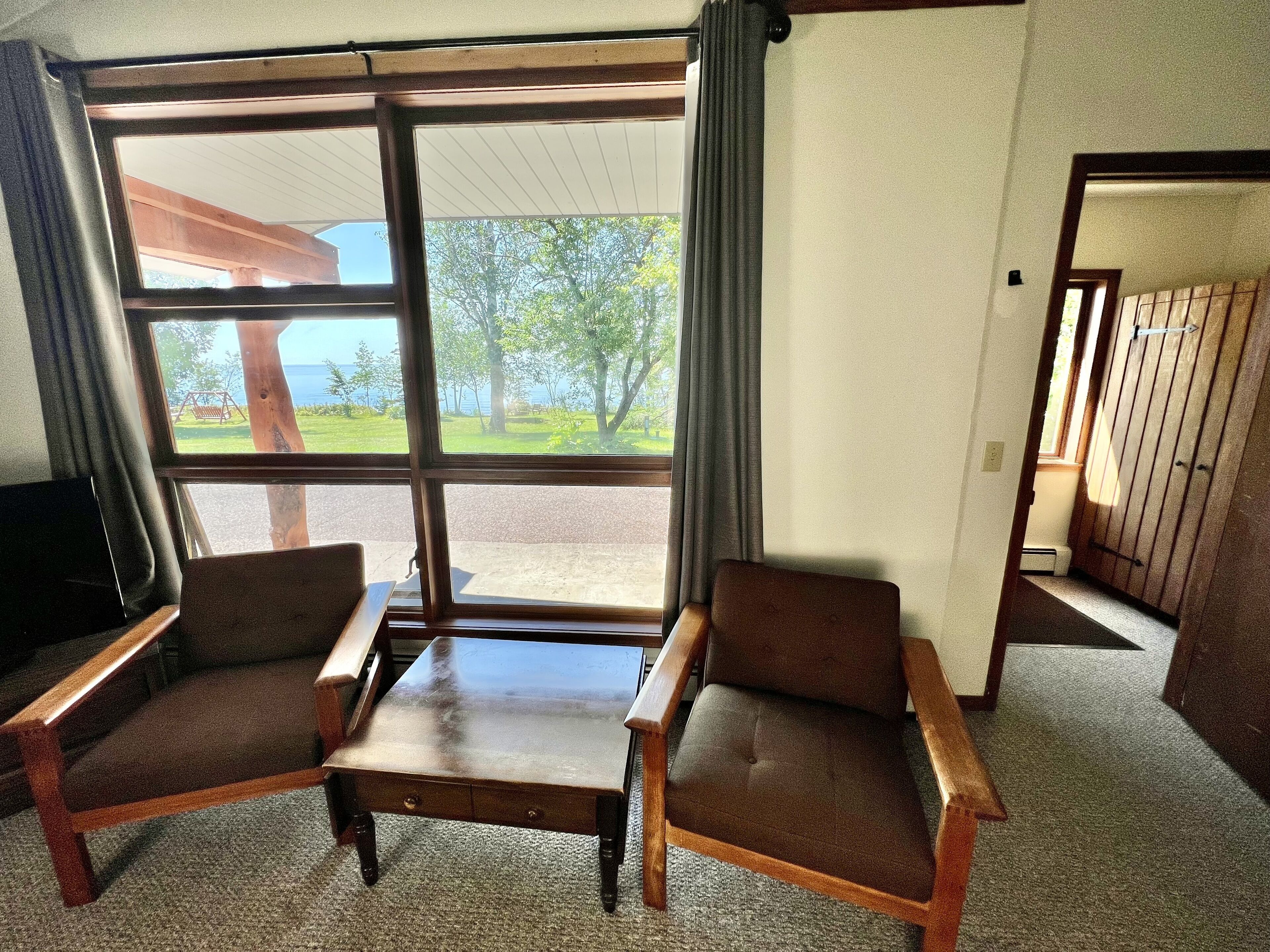 Une pièce avec une grande fenêtre, deux chaises en bois, une petite table en bois et un téléviseur posé sur un meuble.