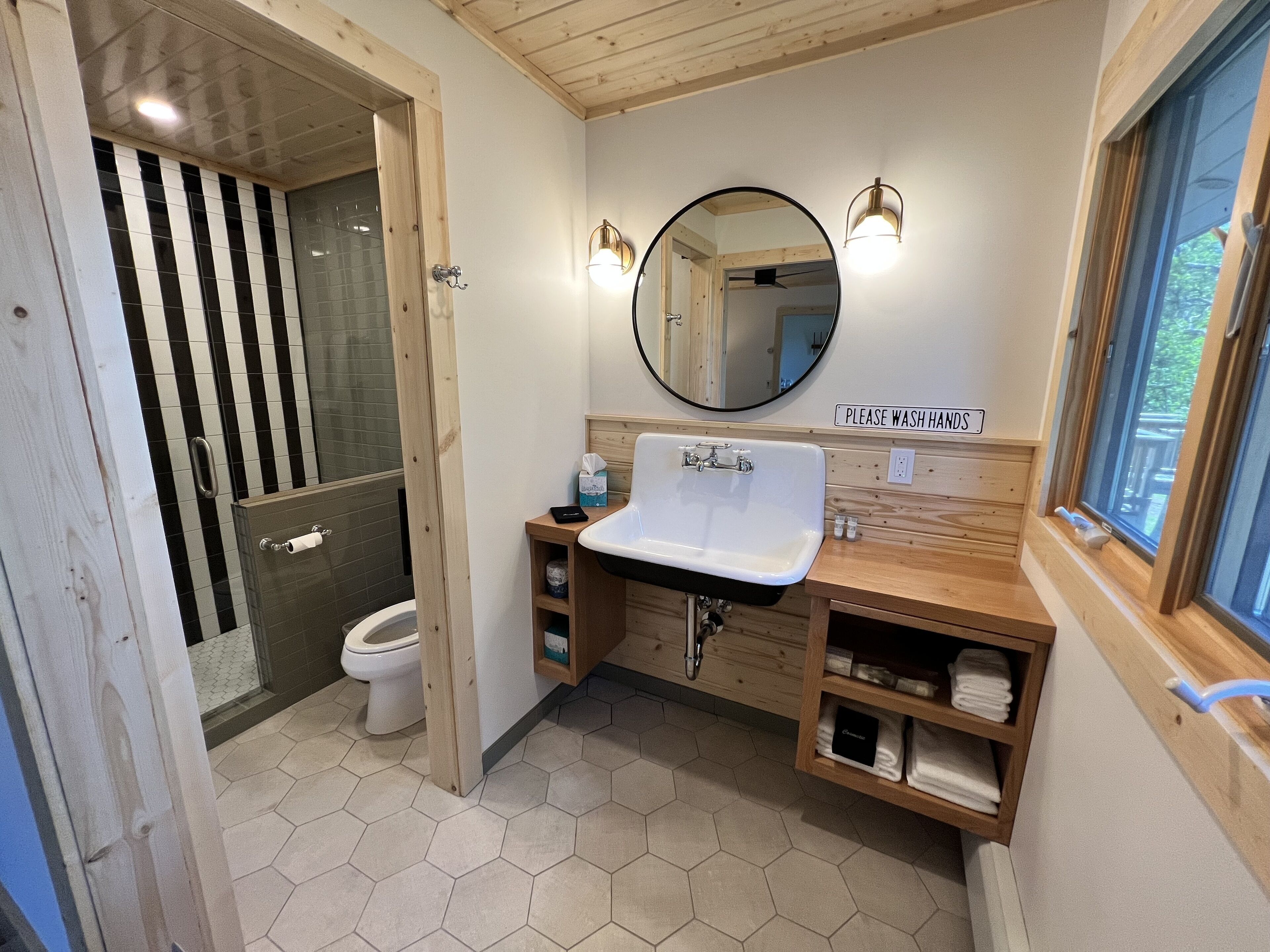 A bathroom with a hexagonal tiled floor, a white sink, wooden cabinets, a mirror, and a sign that says "PLEASE WASH HANDS".