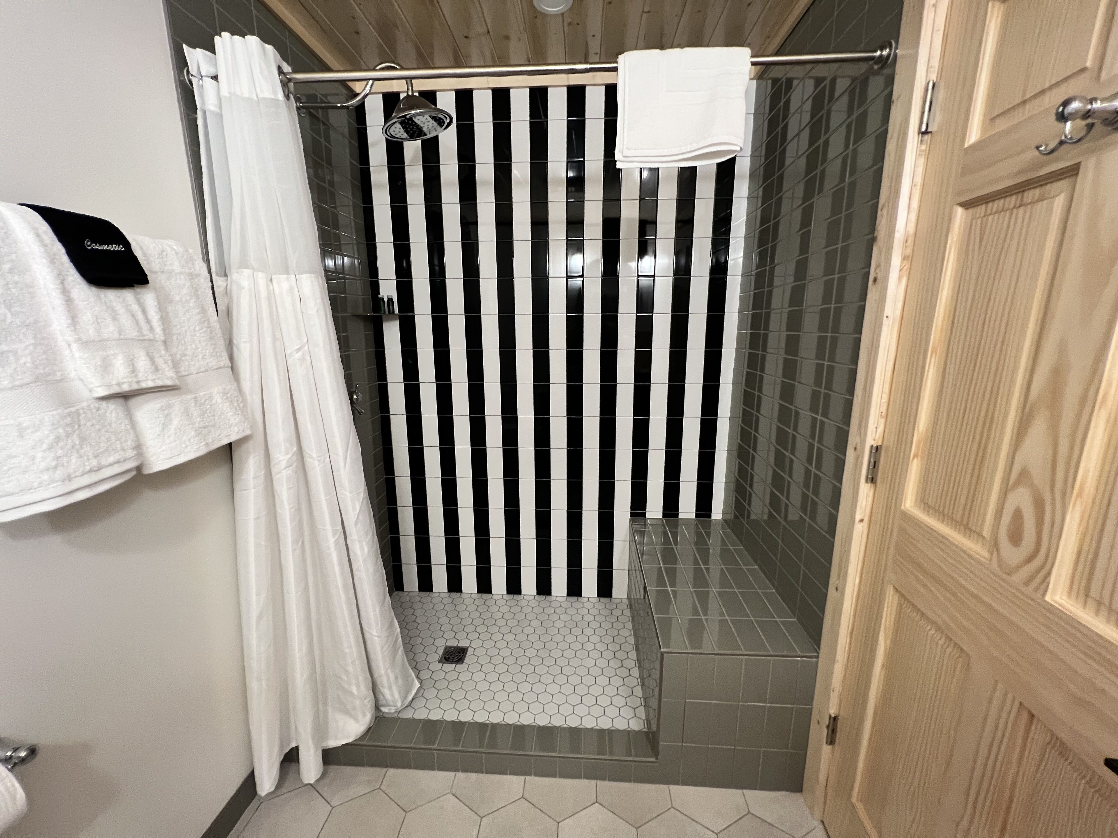 A shower area with a black and white striped shower curtain, a white towel hanging on the wall, and a wooden door.