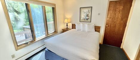A bedroom with a large bed, a nightstand with a lamp, a framed picture on the wall, and a window with a view of a deck and trees.