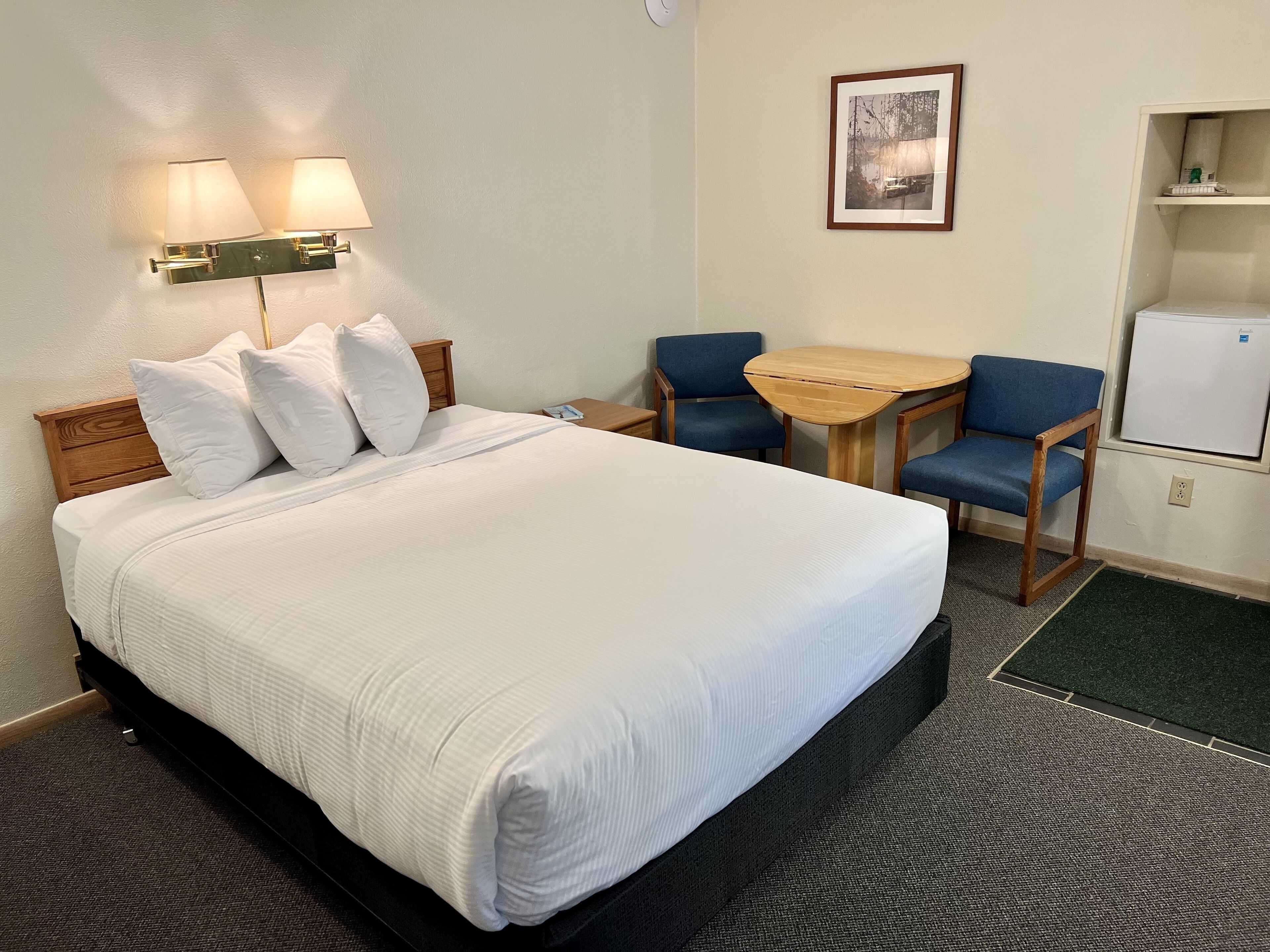 A hotel room with a bed, two chairs, a small table, a wall-mounted lamp, and a refrigerator.