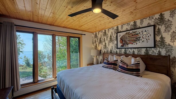 Un dormitorio con una cama grande, un ventilador de techo, un ventanal con vistas al exterior y una pared con papel tapiz con motivos forestales.