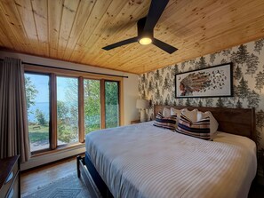 Un dormitorio con una cama grande, un ventilador de techo, un ventanal con vistas al exterior y una pared con papel tapiz con motivos forestales.