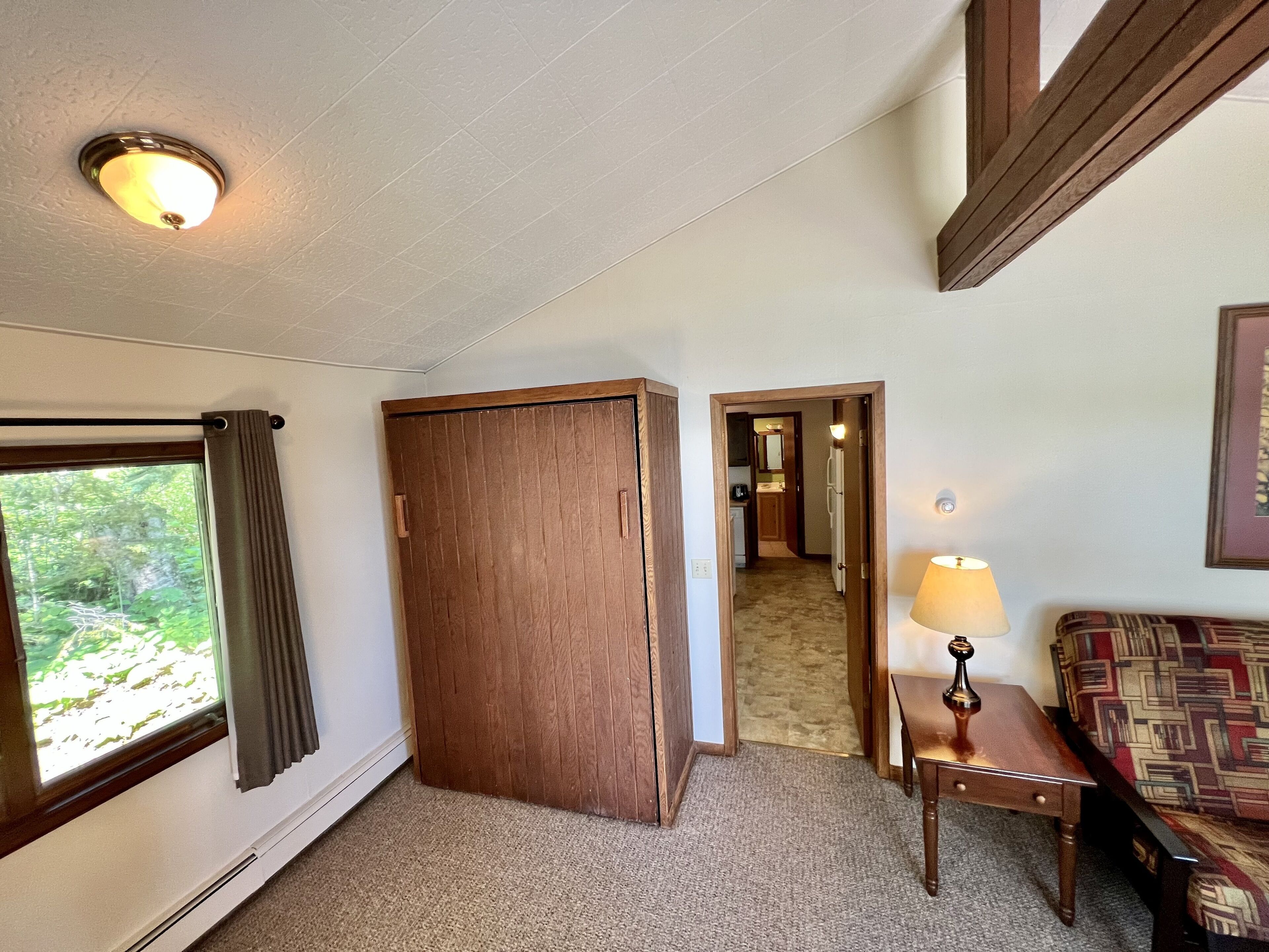A room with a wooden wardrobe, a small table with a lamp, a patterned sofa, and a window with curtains.