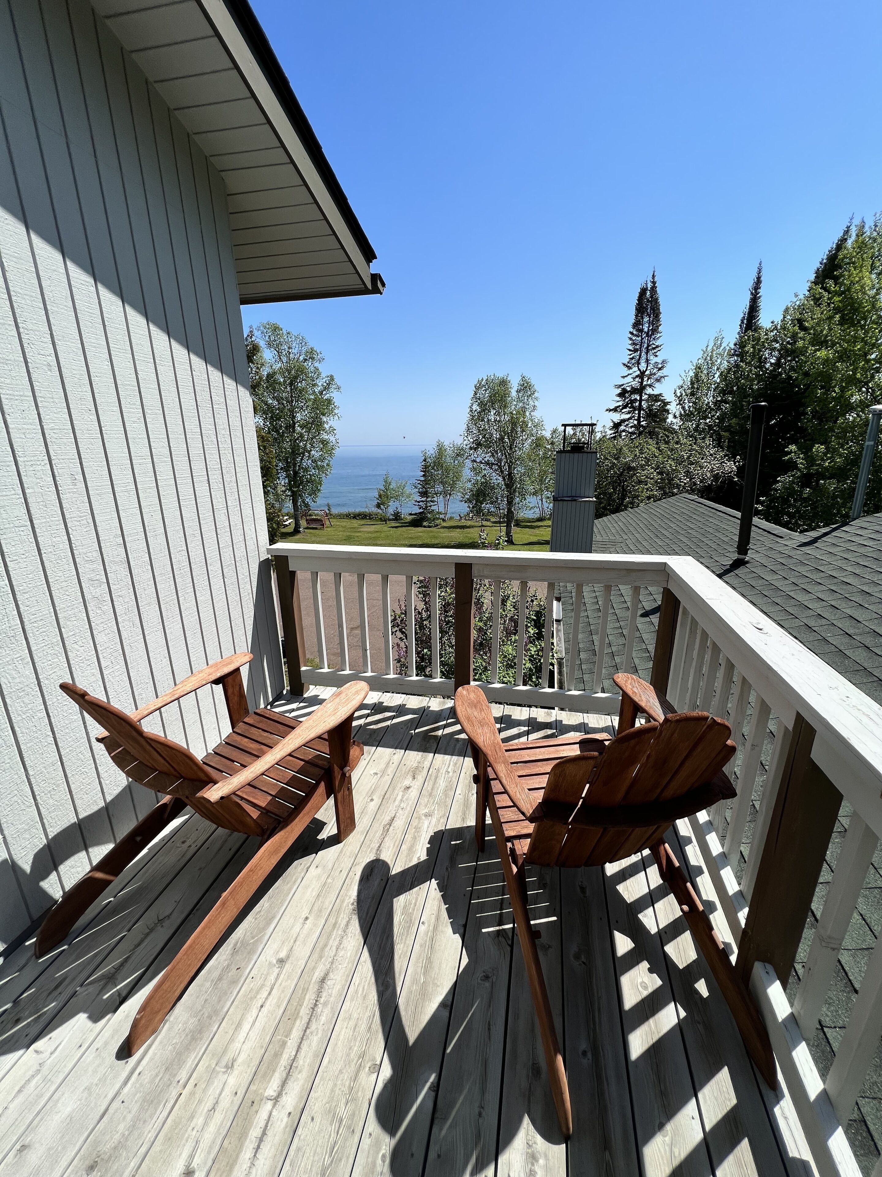 Um deck de madeira com duas cadeiras Adirondack voltadas para uma vista panorâmica de um corpo d'água e árvores.