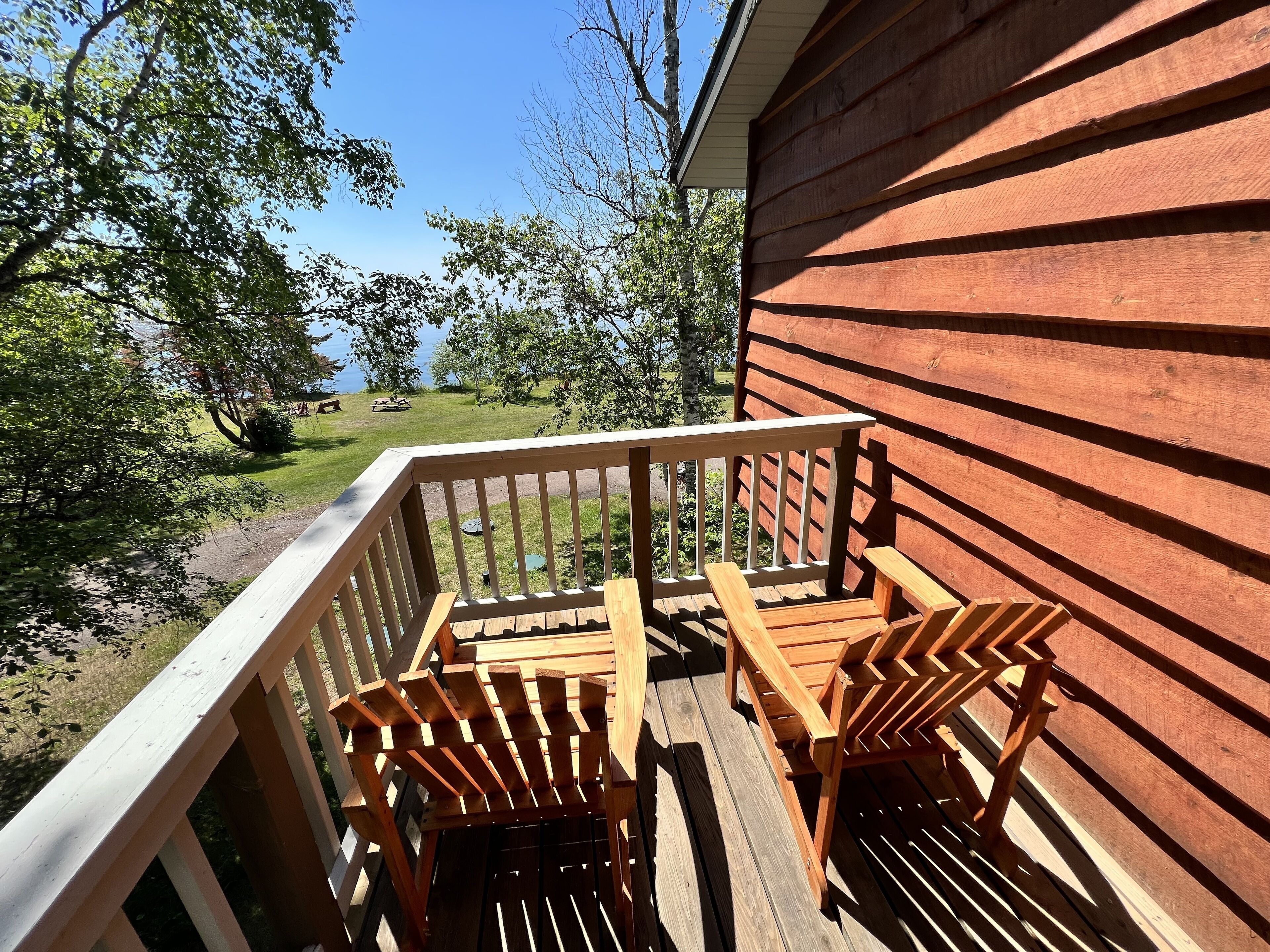 A wooden deck with two Adirondack chairs and a table, overlooking a grassy area with trees and a body of water.
