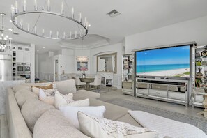 Ein geräumiges Wohnzimmer mit einem großen Flachbildfernseher, der eine Strandansicht zeigt, einer modernen Kronleuchter und einer gemütlichen Sitzecke.