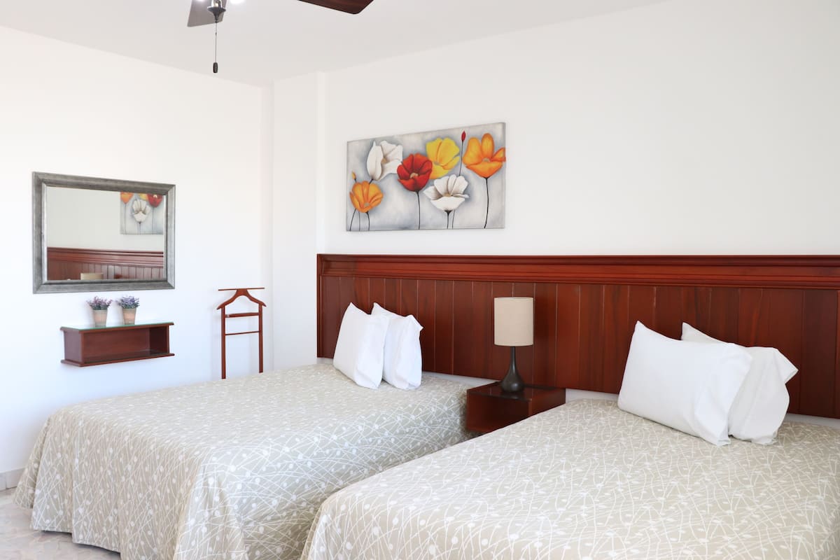 A hotel room with two beds, a wooden headboard, a nightstand with a lamp, a wall-mounted mirror, and a floral painting.
