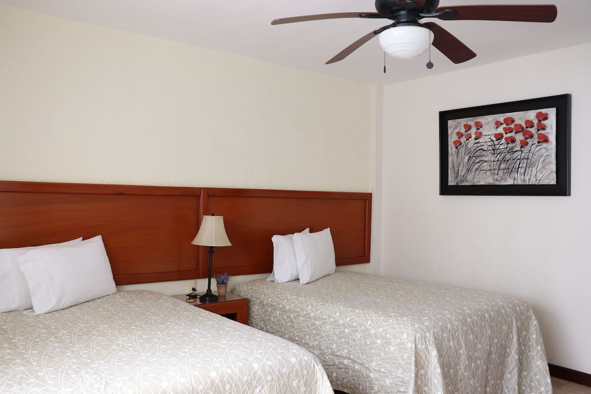 A hotel room with two beds, a wooden headboard, a ceiling fan, a nightstand with a lamp, and a framed artwork on the wall.