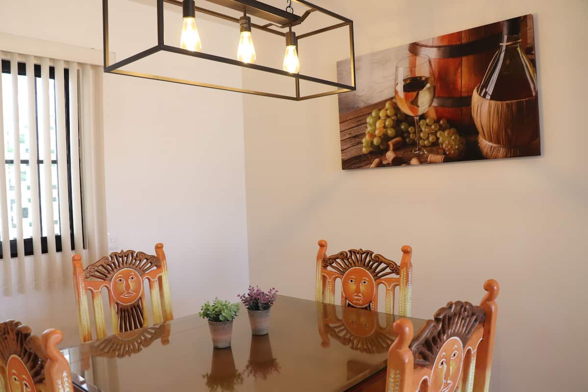 A dining area with a glass table, wooden chairs, a chandelier, and a wall-mounted painting of grapes and a wine bottle.