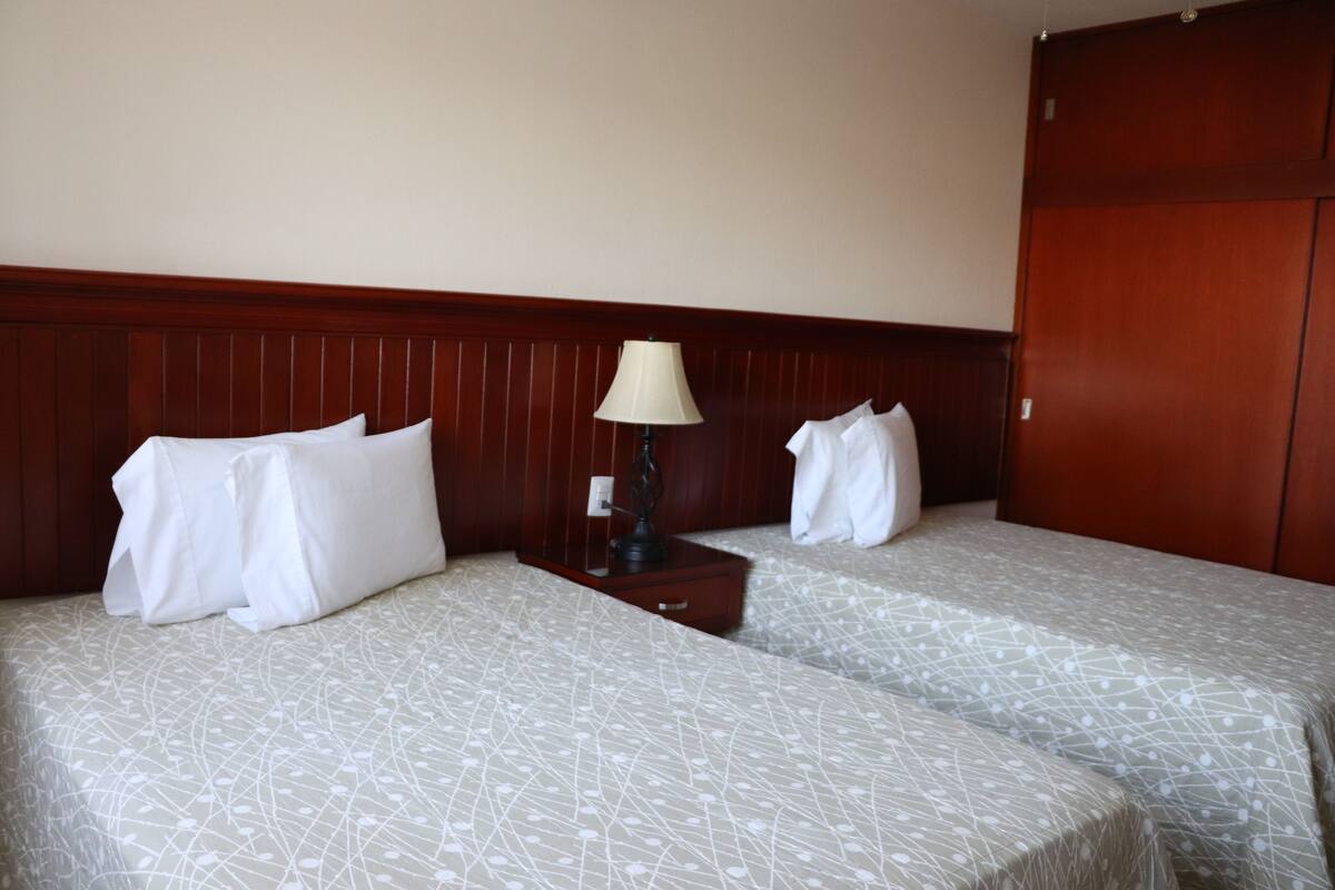 A hotel room with two beds, a wooden headboard, a bedside table with a lamp, and a wardrobe.