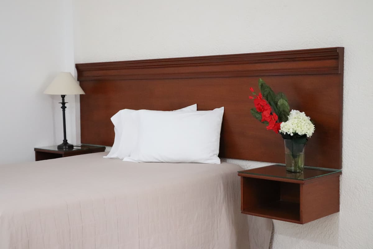 A neatly made bed with white pillows and a beige bedspread, a bedside table with a lamp, and a small wooden shelf with a vase of flowers.