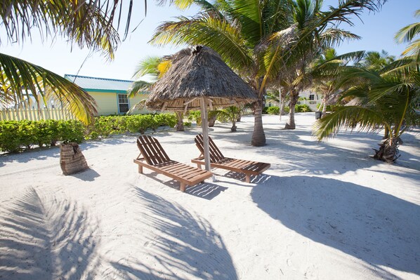 A beach area with two wooden lounge chairs under a thatched umbrella, surrounded by palm trees and a building with a blue roof.