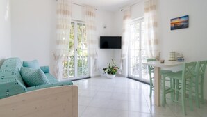 Un salón luminoso con un sofá, una mesa de comedor con sillas, un televisor montado en la pared y un balcón con cortinas.
