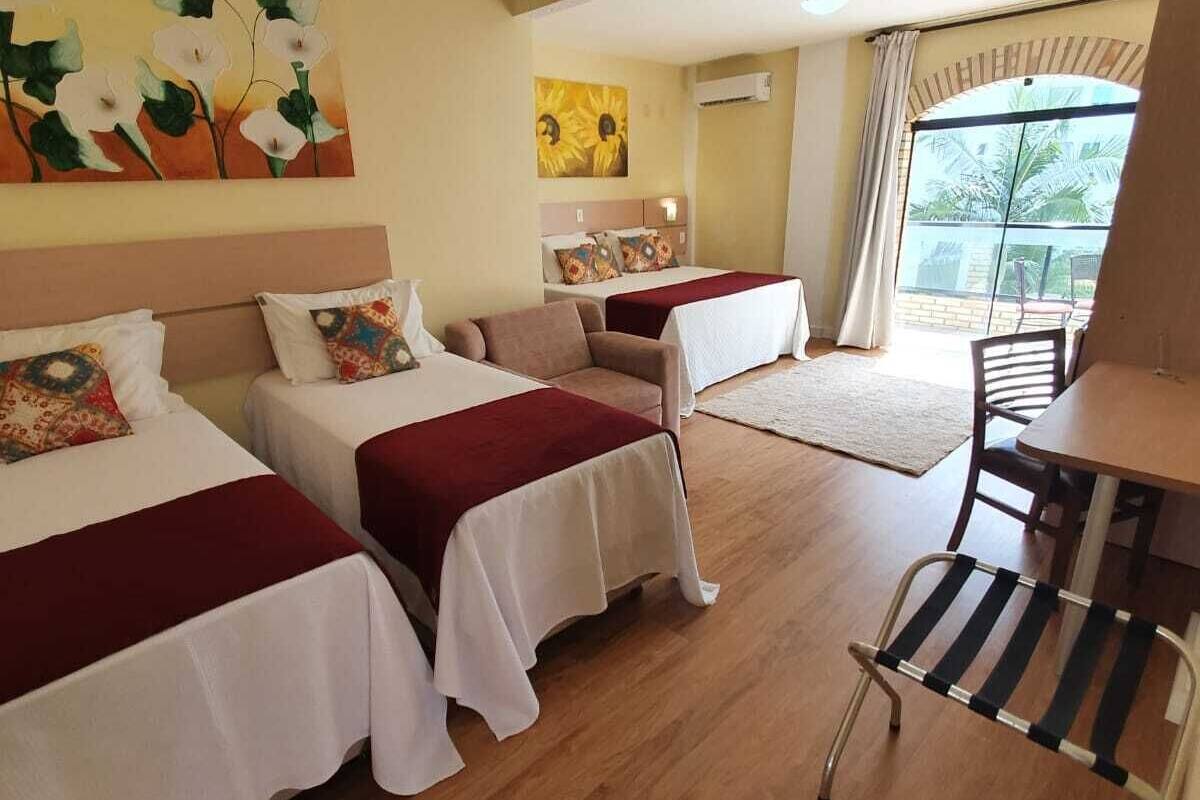 A hotel room with two beds, a dining table, and a chair. There are paintings on the walls and a large window with curtains.