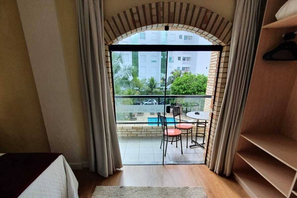 A room with a bed, a wooden floor, a large window with curtains, a view of a balcony with outdoor furniture, and a small table.