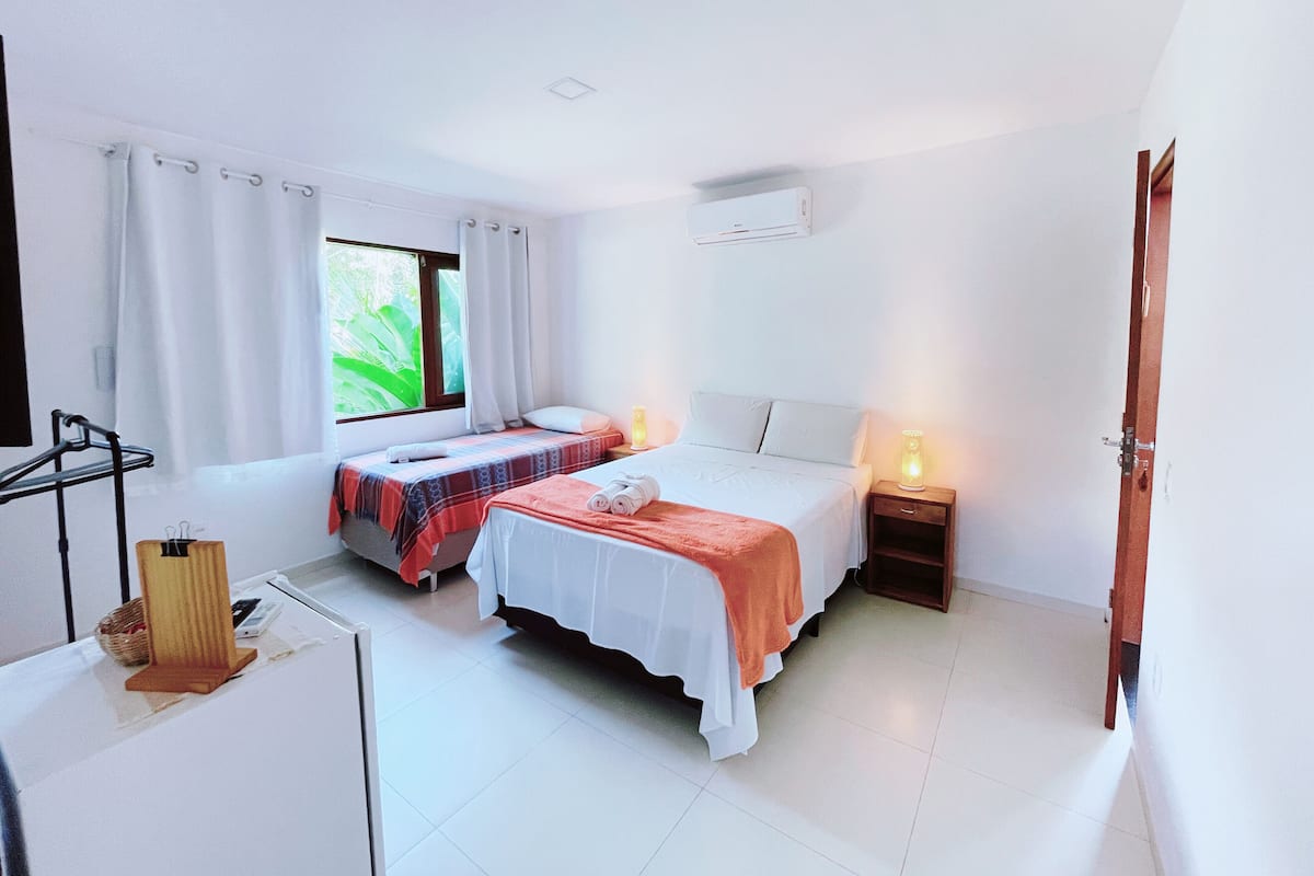 A hotel room with two beds, a window with curtains, a bedside table with a lamp, and a small table with a basket and a phone.