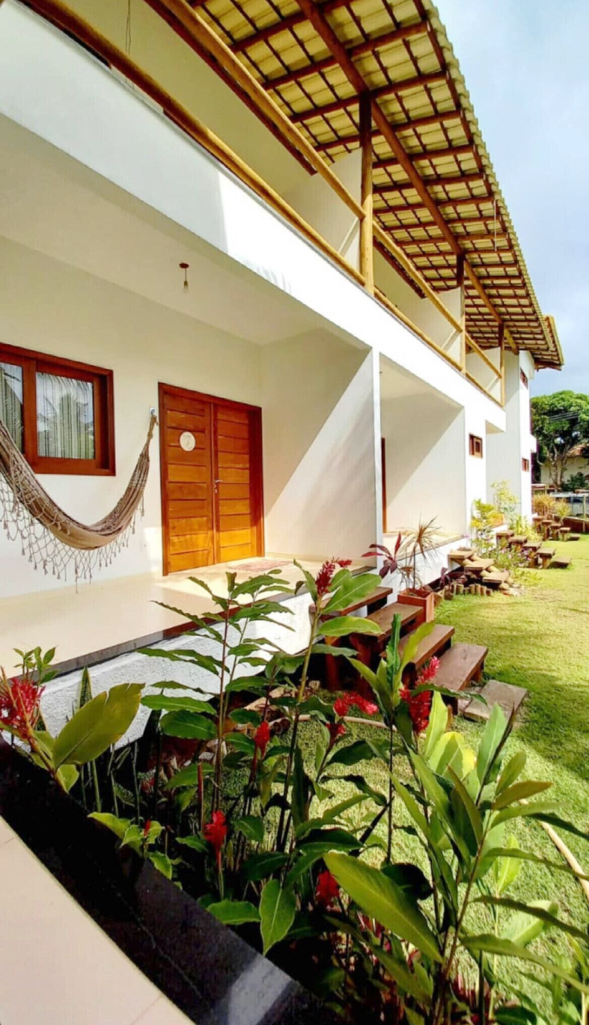 Uma casa moderna com varanda coberta, uma rede e um jardim bem cuidado com diversas plantas e flores.