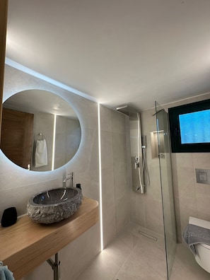 A modern bathroom with a round mirror, a stone sink, a glass shower enclosure, and a toilet.