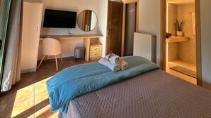 Ein Schlafzimmer mit Holzbalkendecke, einem Bett mit blau-brauner Bettwäsche, einem Schreibtisch mit Computer, einem Stuhl und einer kleinen Pflanze.