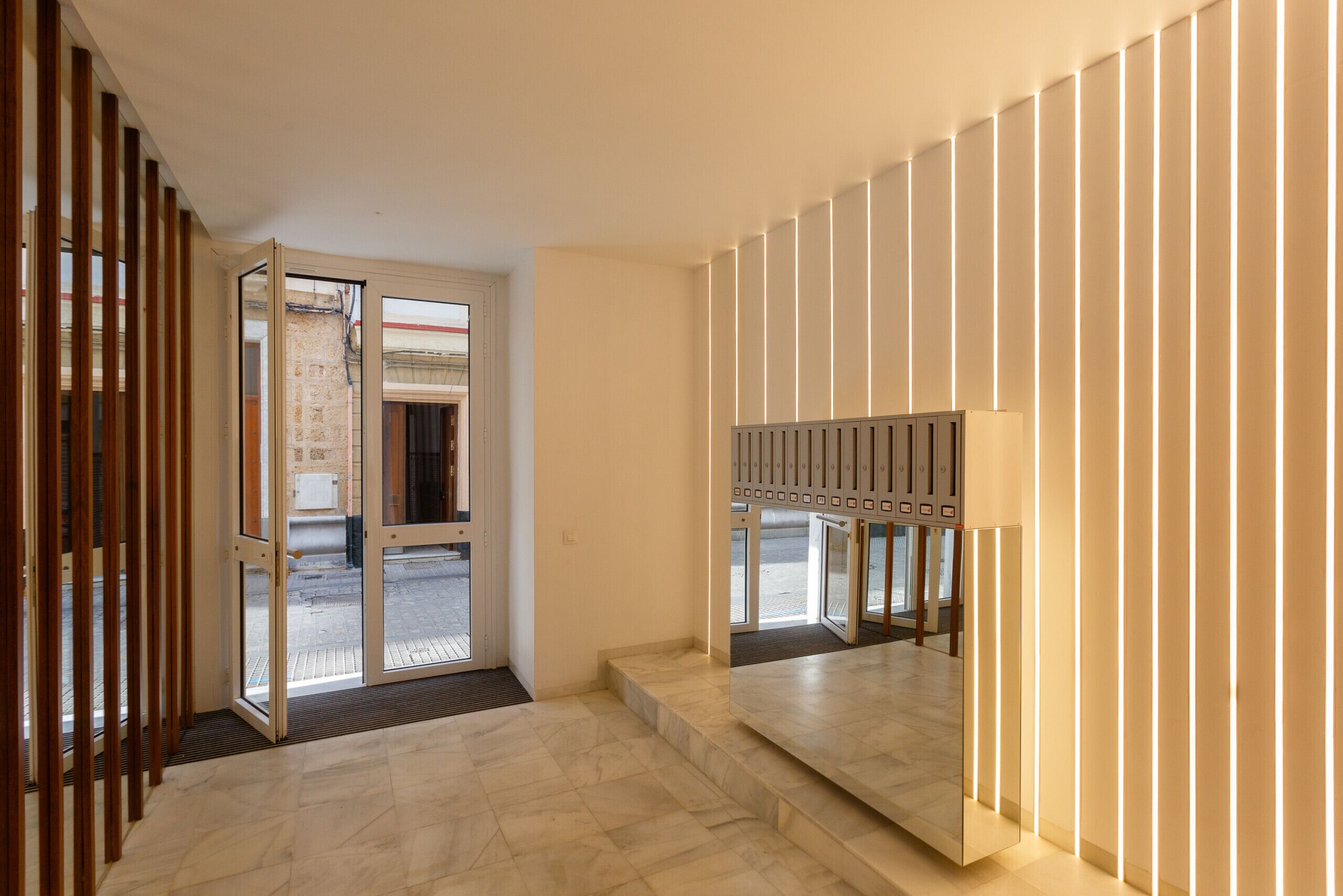 Un interior moderno con un piso de mármol, una puerta de cristal que da al exterior y una pared con paneles de luz verticales.
