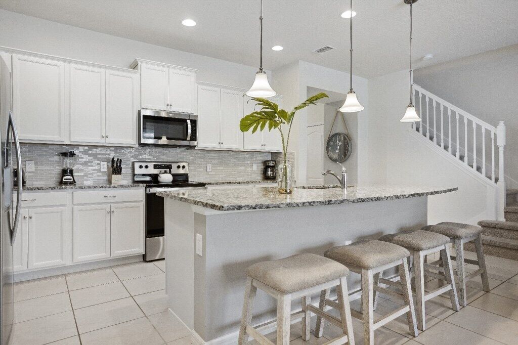 Una cocina moderna con armarios blancos, una isla de granito, taburetes de bar e iluminación colgante.