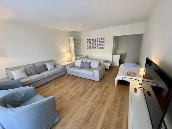 Un salon moderne avec un canapé gris, un fauteuil blanc, une petite table blanche et un lit avec un téléviseur.