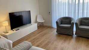 A modern living room with a flat-screen TV on a white cabinet, two grey armchairs, a white chair, and a lamp.